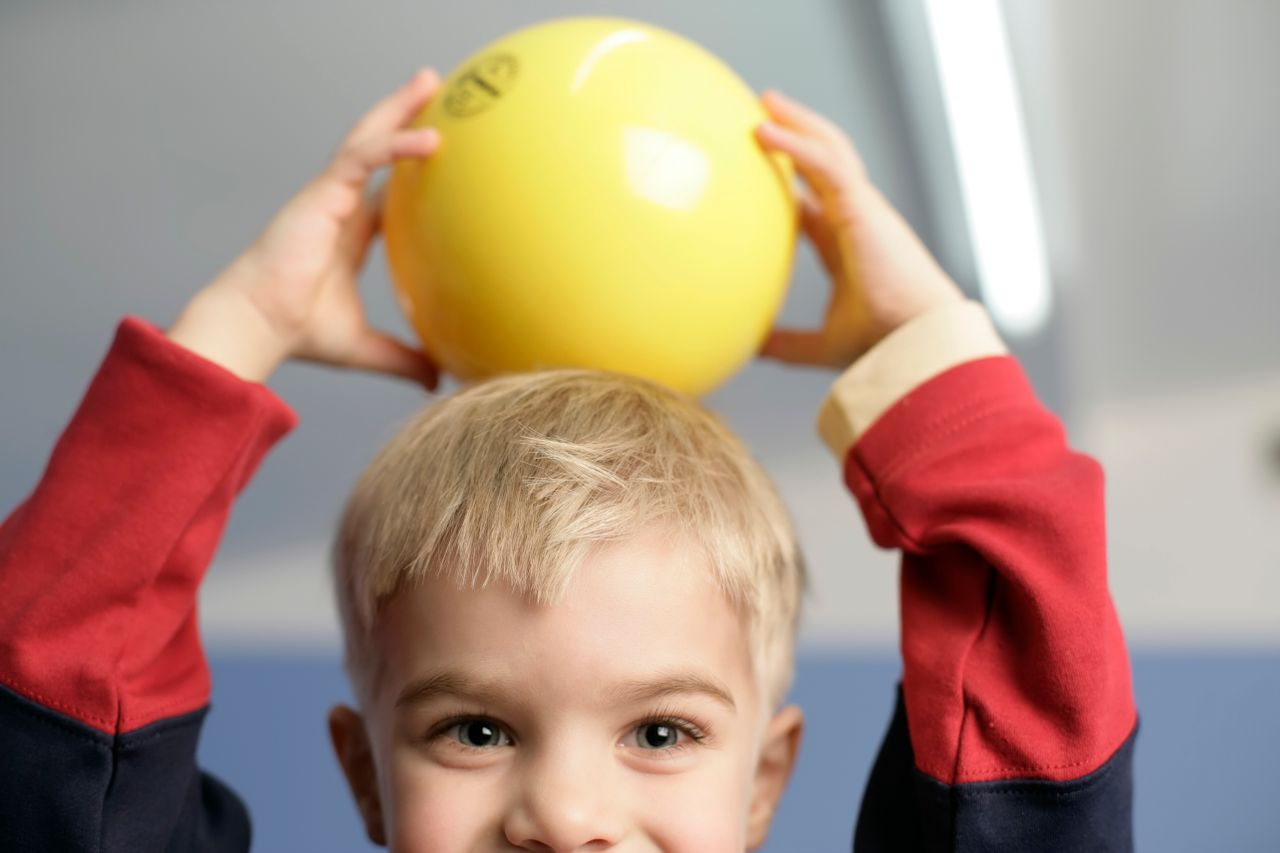 A cheerful child holding a yellow ball above their head, indoors.